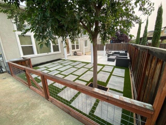 Backyard patio with stone squares, artificial grass, tree, wooden fence, and black outdoor furniture.