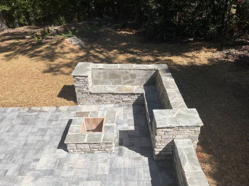 Stone patio with built-in seating and a fire pit in a yard with gravel and trees.
