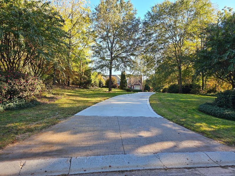 Concrete driveway curves toward a house, surrounded by green grass and trees on a sunny day.