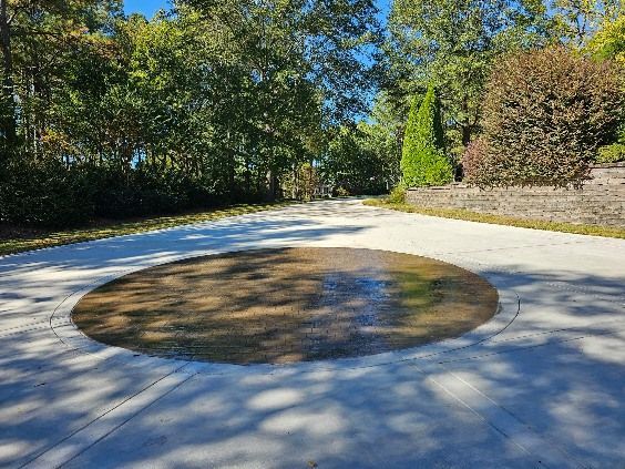 Circular area of wet dirt on a concrete driveway with trees in the background. Sunny day.