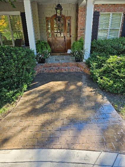 Brick-patterned walkway leading to a house with a brown door flanked by bushes and potted plants.