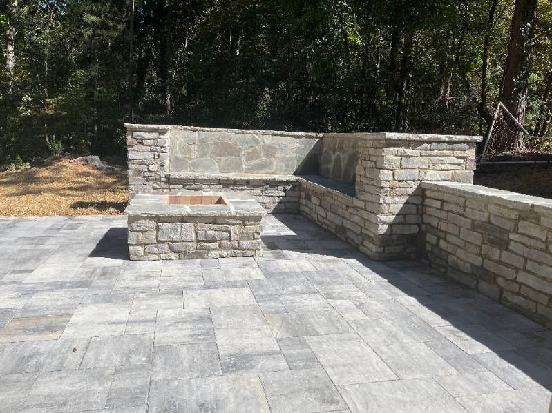 Stone patio with built-in bench and fire pit, set against a wooded backdrop.