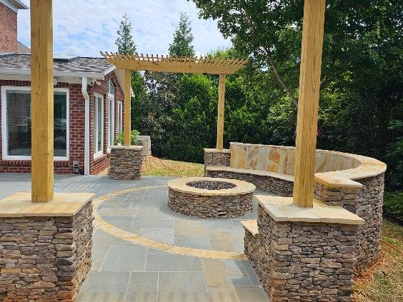 Stone patio with built-in bench, fire pit, and pergola, set against a house and trees.