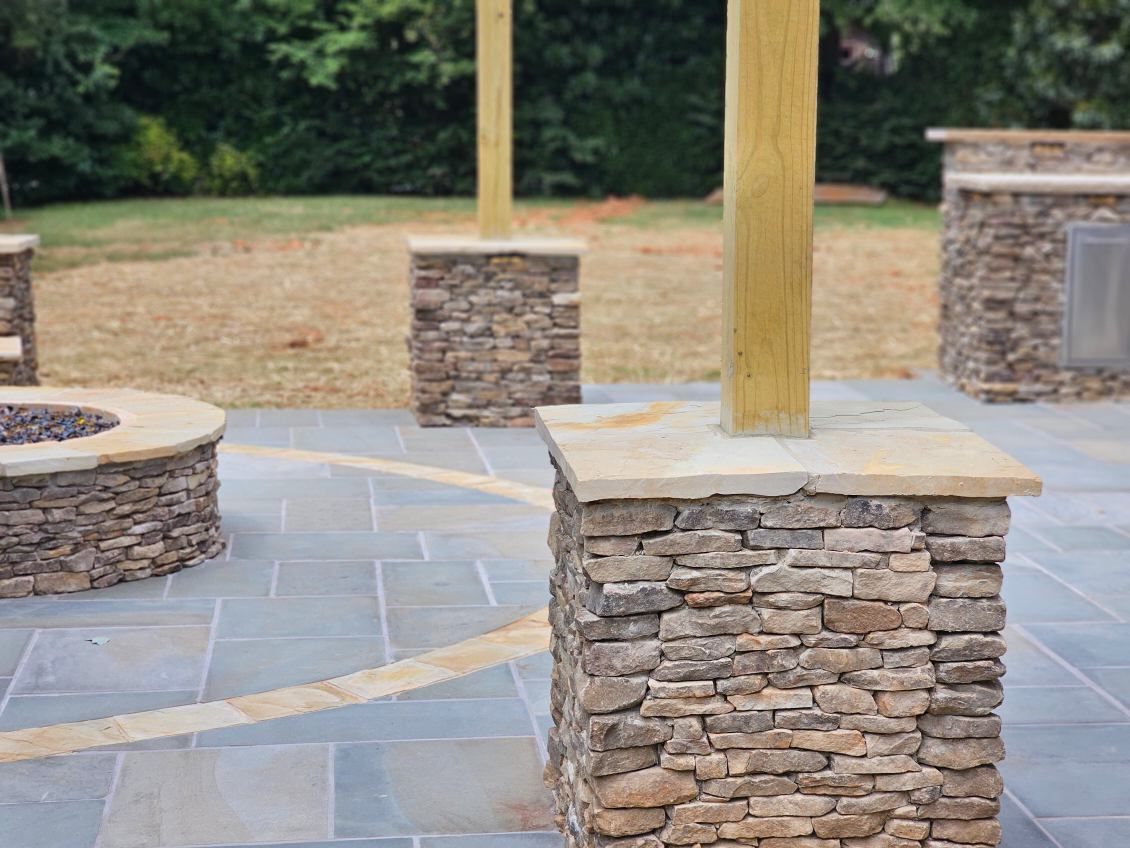 Stone pillars with wooden posts on a paved patio, with a fire pit and grill in the background.