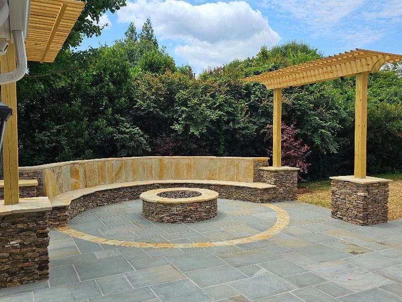 Outdoor stone patio with fire pit, curved seating, and wooden pergola under a blue sky.