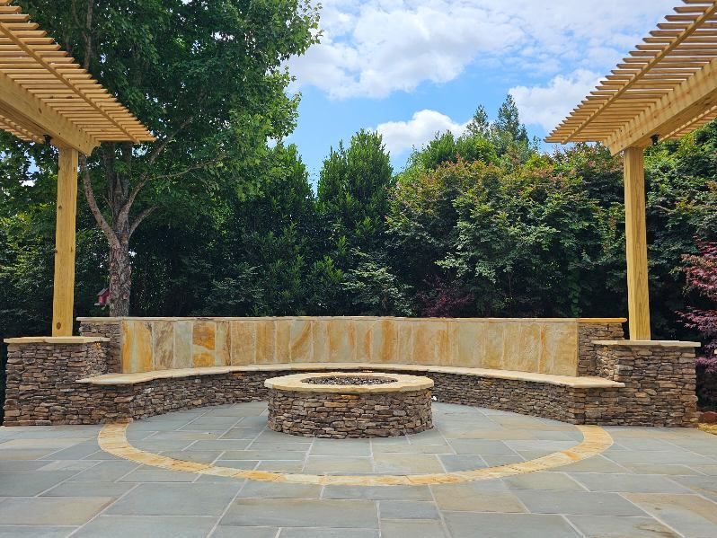Outdoor fire pit area with stone seating, pergola, and lush greenery.