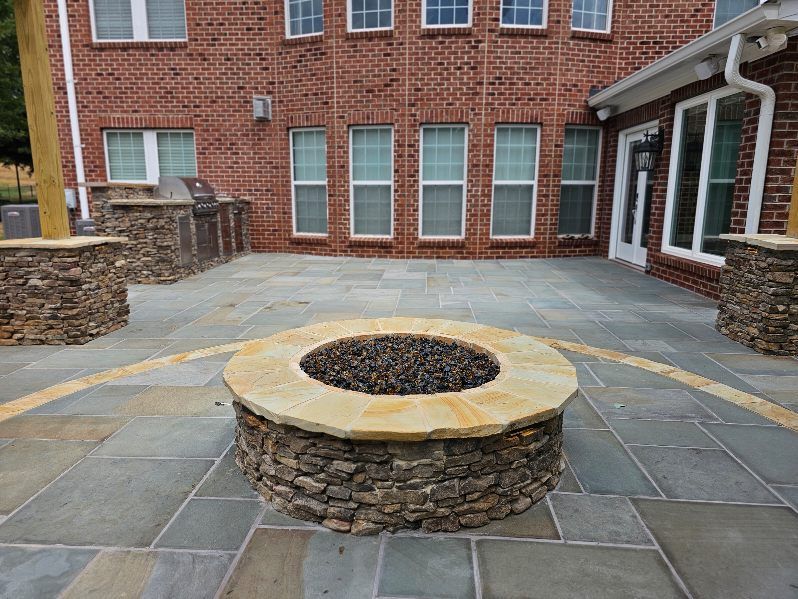 Stone fire pit on a blue-gray patio. Red brick building with windows in background.