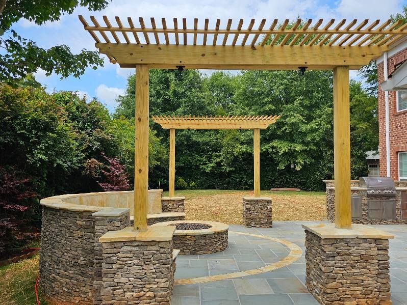 Outdoor patio with stone walls, wooden pergola, and fire pit under a blue sky.