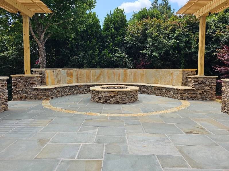 Stone patio with fire pit, built-in curved seating, and pergola posts under a blue sky.