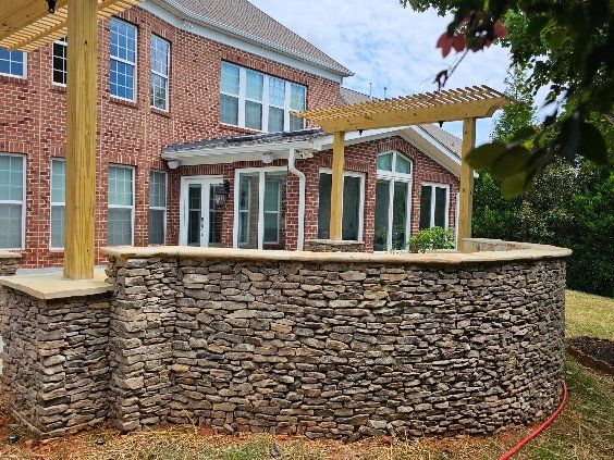 Stone-walled patio with pergola against a brick house. Green lawn surrounds the structure.