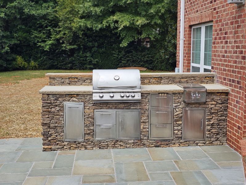 Outdoor stone kitchen with grill, cabinets, and built-in toaster, next to a brick wall.