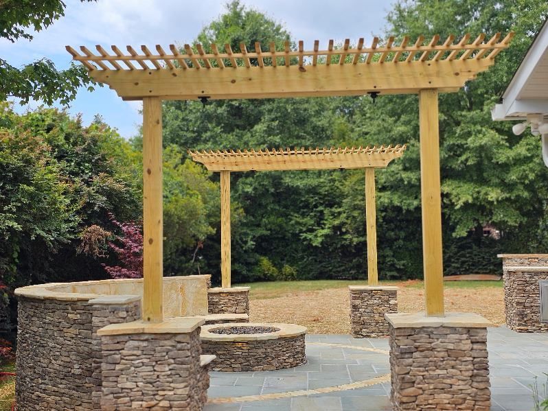 Pergolas with stone bases over a fire pit and seating area in a backyard.