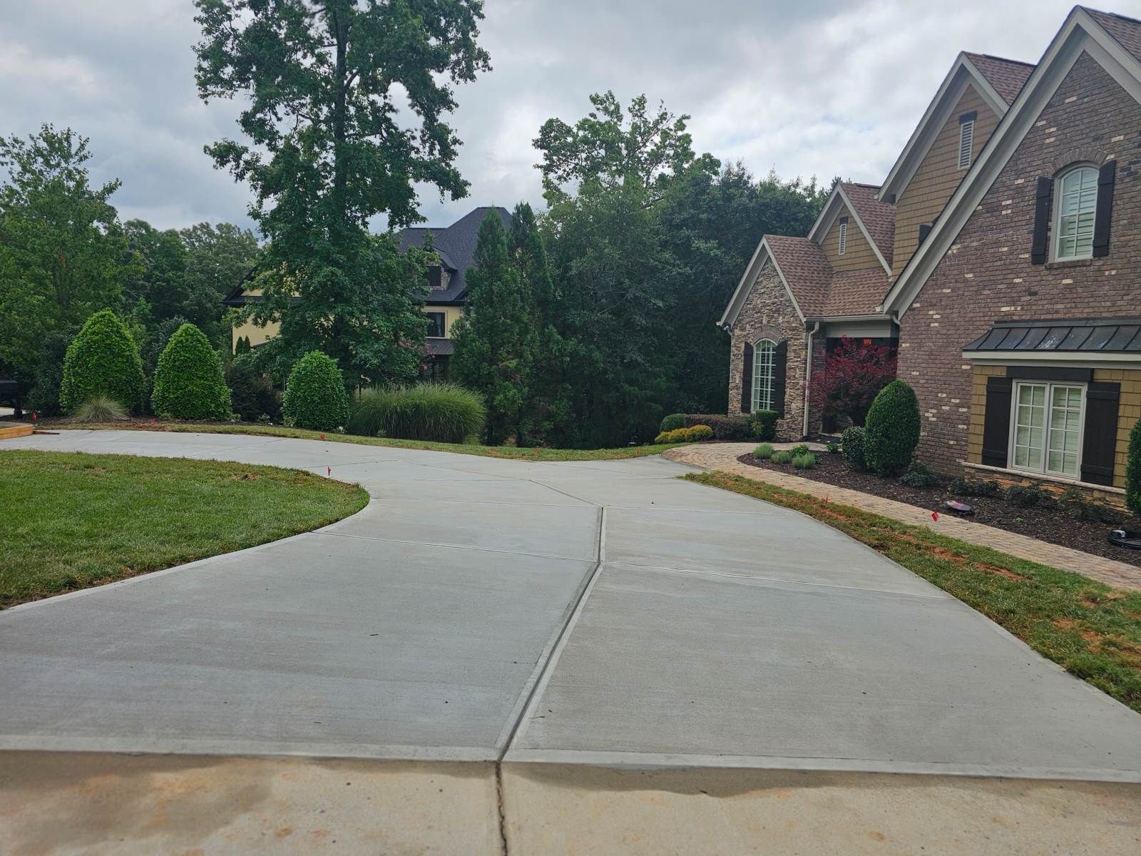 Driveway leading to a brick house on a cloudy day, with trees and a lawn.