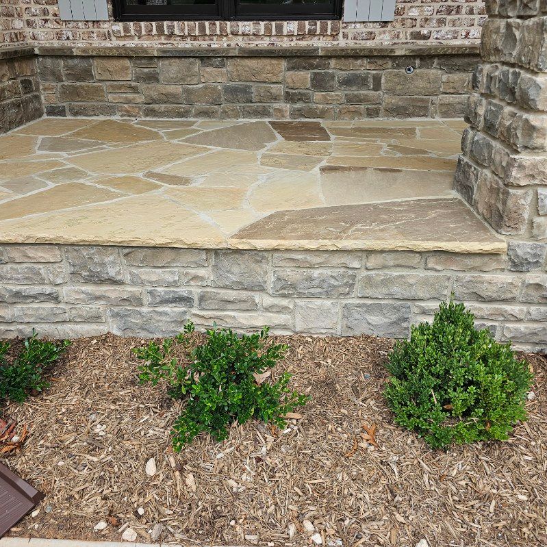 Stone porch with irregular flagstone flooring and stacked stone facade, small shrubs in mulch.