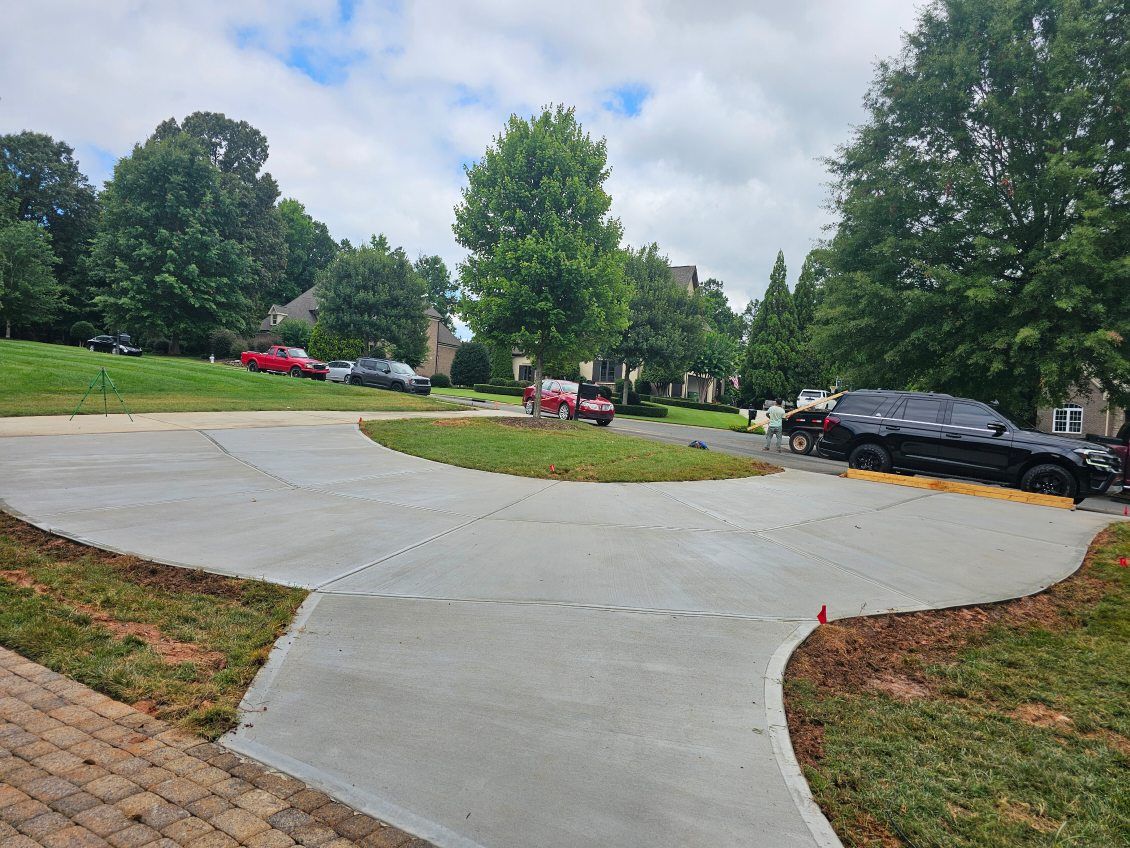 Concrete driveway intersection with grassy median, trees, parked cars, cloudy sky.