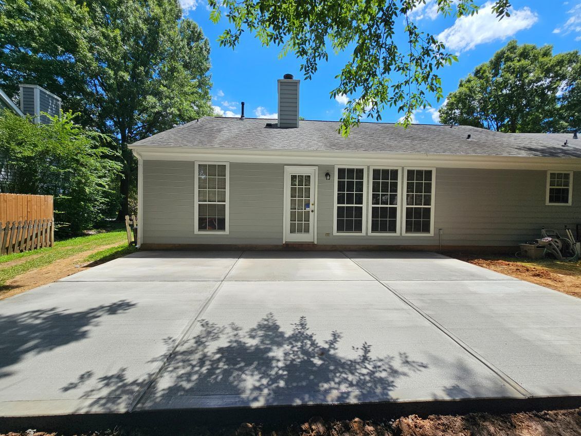 Back of a light gray house with a new concrete patio, trees and blue sky.
