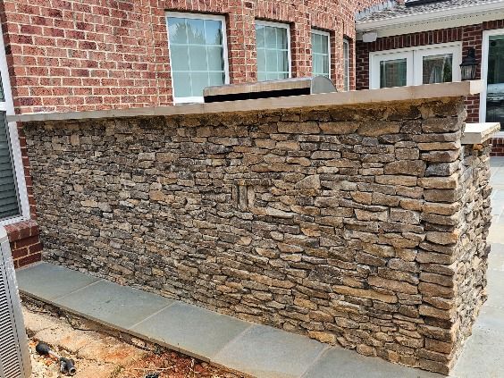 Stone-covered outdoor kitchen counter with brick house background and a concrete base.