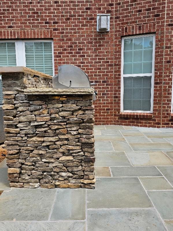 Outdoor kitchen island with stone facade and grill, against a brick wall with windows, on a stone patio.