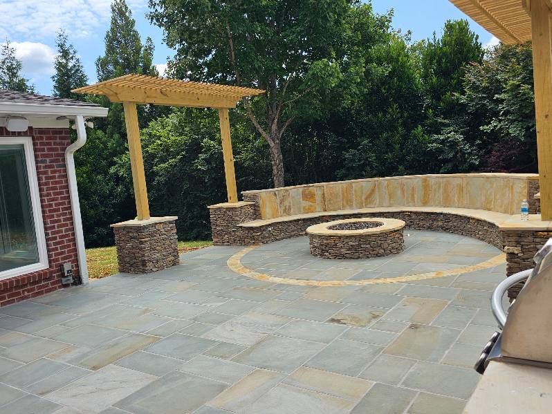 Stone patio with built-in seating, a fire pit, and a wooden pergola, surrounded by trees.