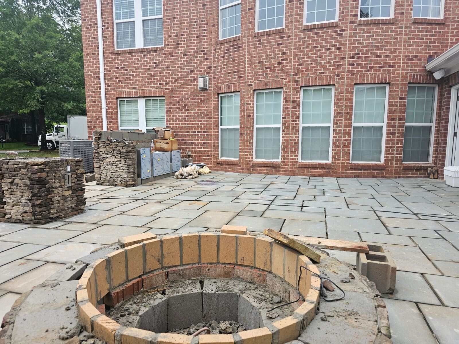 Brick fire pit under construction on a stone patio in front of a brick building.