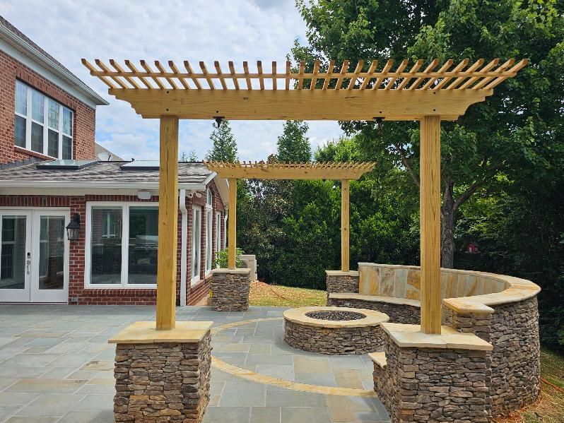 Wooden pergola over stone patio with built-in seating and fire pit, next to a brick house.