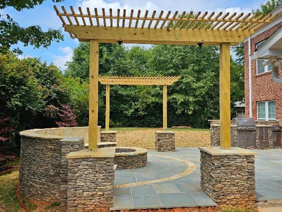 Stone patio with wooden pergola, circular fire pit, and brick house. Blue sky and green trees in background.