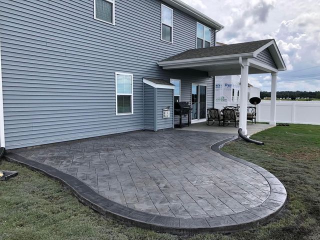 Gray stamped concrete patio next to a light gray house. Curved border, grass, and a covered porch.