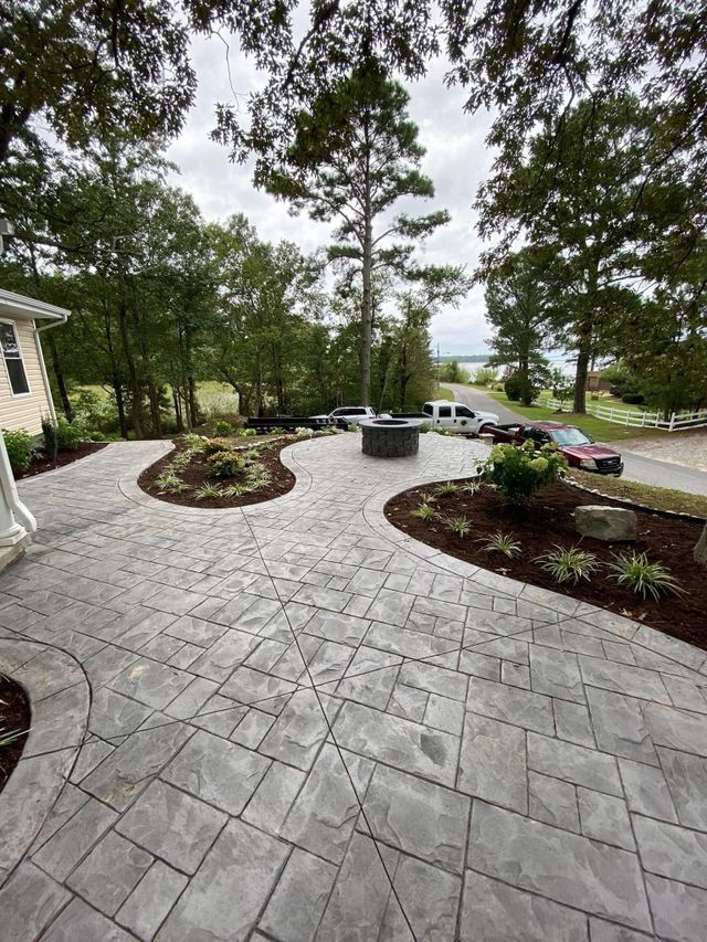 Gray paved patio with circular planting beds, a fire pit, and trees in the background.