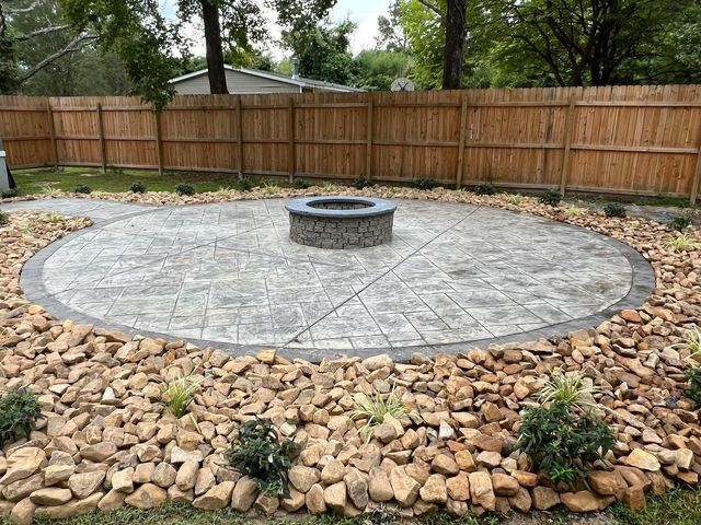 Circular concrete patio with fire pit, bordered by rocks and landscaping, and wooden fence.
