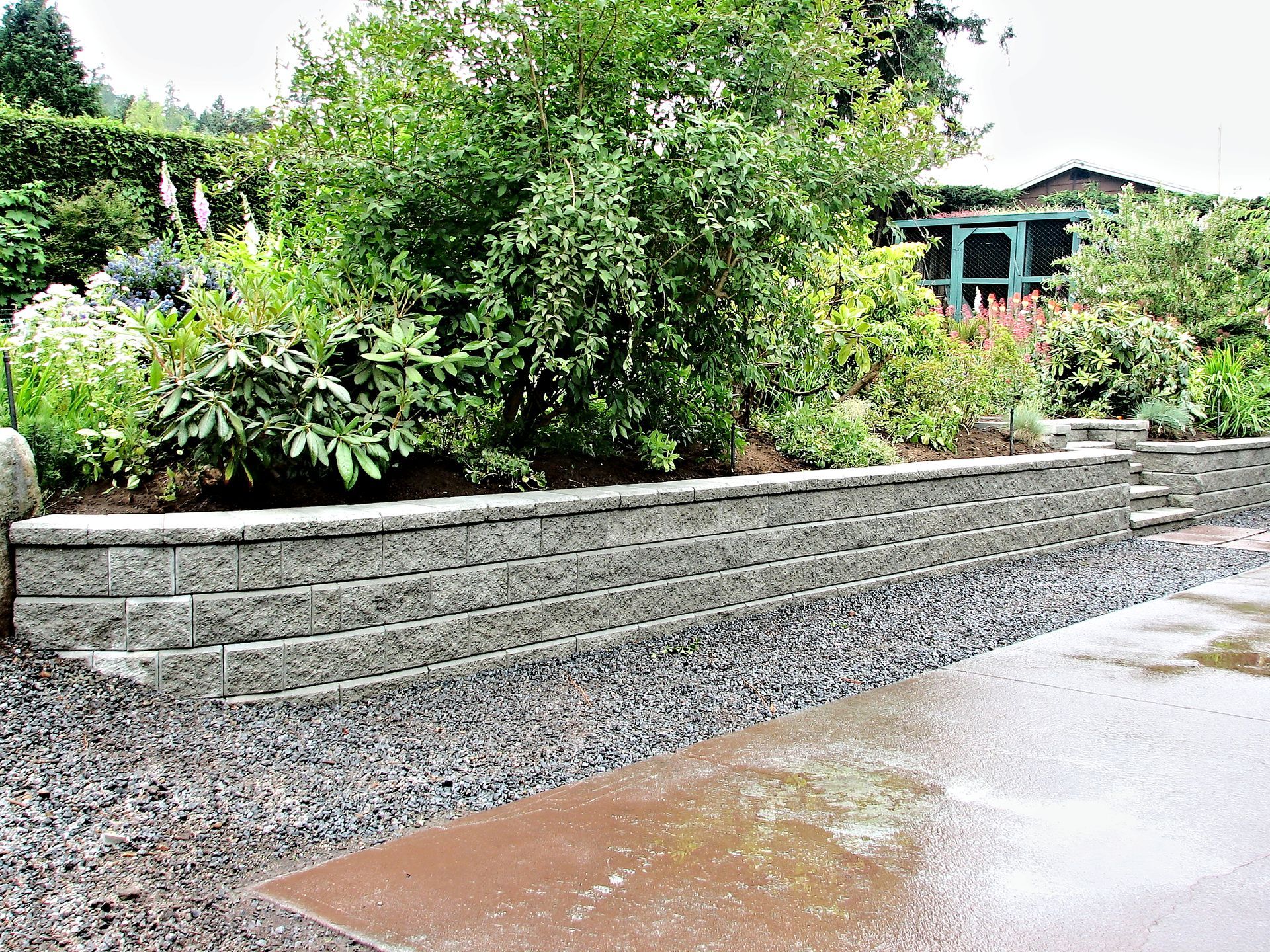 Retaining wall made of gray blocks with gravel, plants, and a small green structure in the background.