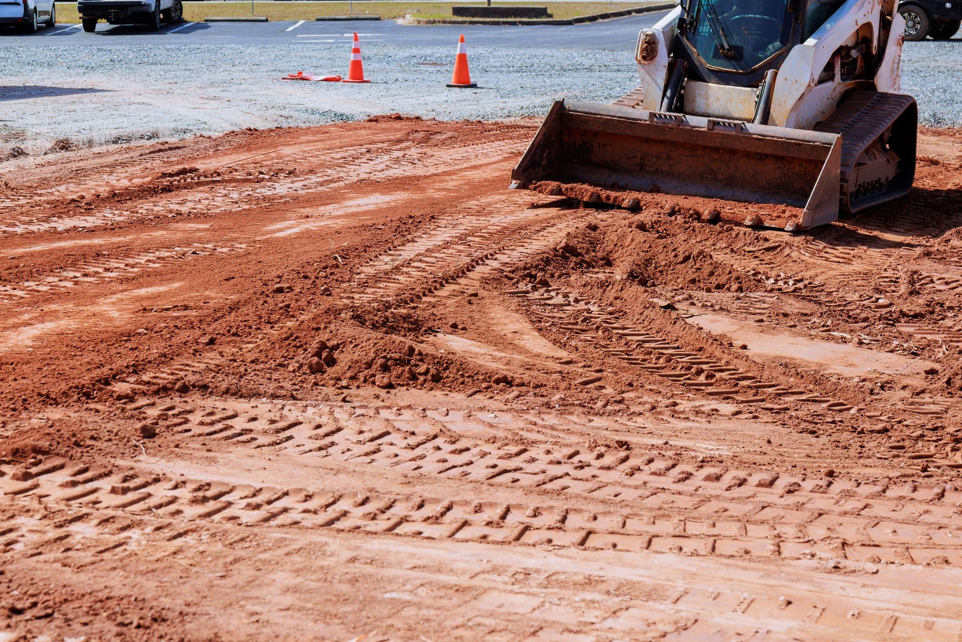 Skid steer on a red clay surface, making tracks. Construction cones in background.