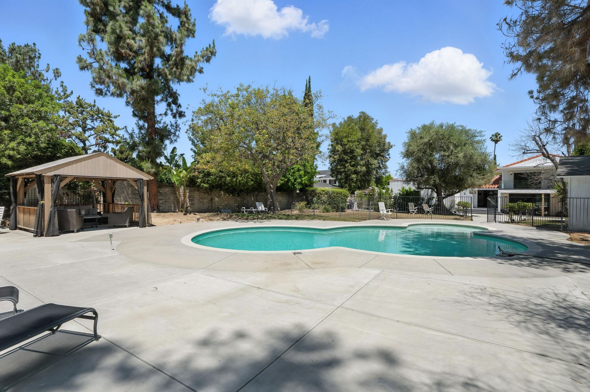 A backyard pool with a gazebo, trees, and lounge chairs on a sunny day.