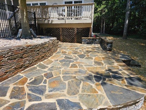 Flagstone patio leading to a wooden deck, bordered by a stone retaining wall and grass.