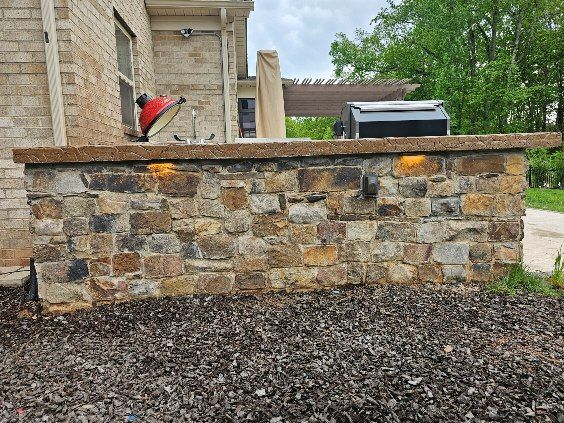 Stone outdoor kitchen with grill and counter, lit by warm lights. Brown and tan stonework with mulch below.