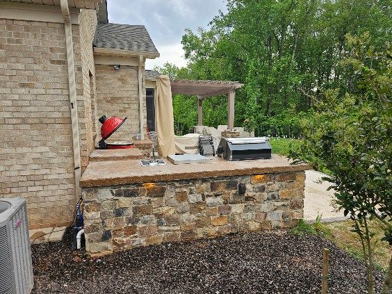 Stone patio with a grill and overhead pergola, next to a brick house.