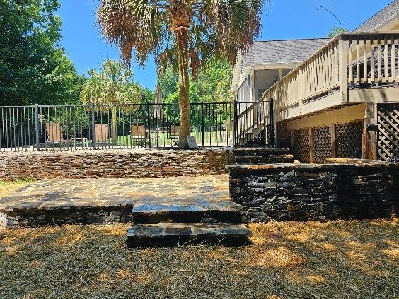 Backyard with stone retaining walls, steps, black metal fence, wooden deck, and palm tree on a sunny day.