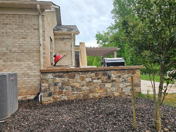 Outdoor stone kitchen with grill, brick house, and pergola in a backyard.