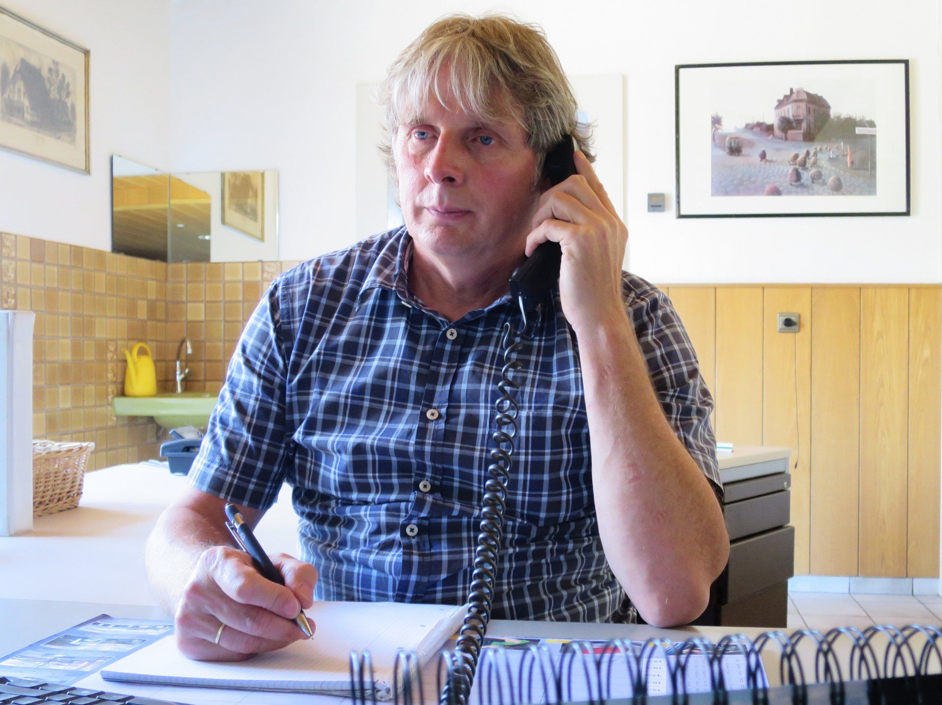 Eine Person in einem blau karierten Hemd sitzt an einem Schreibtisch, telefoniert mit einem Festnetztelefon und schreibt 