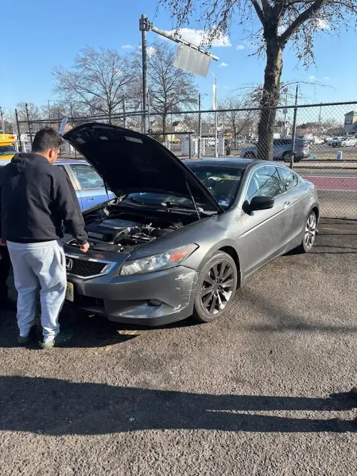 Man looking at a gray car with its hood open; damage to the front. Outside on a sunny day.