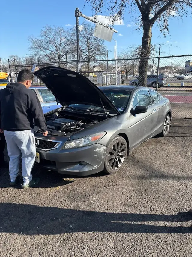 Man looking at a gray car with its hood open; damage to the front. Outside on a sunny day.