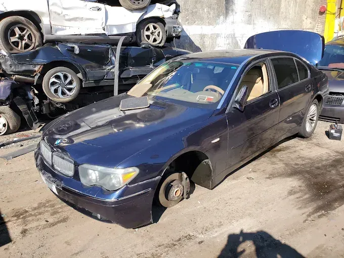 Dark blue BMW sedan in a salvage yard, missing wheels. Other wrecked cars visible.