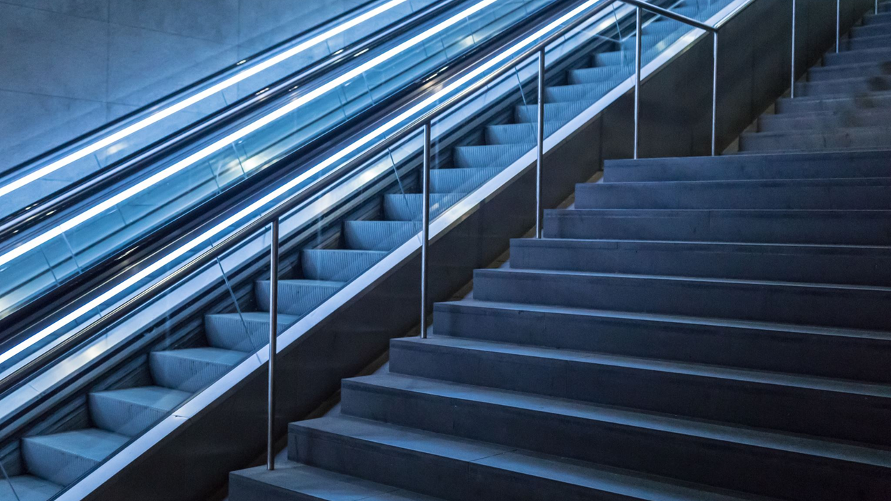 Escalator and stairs side by side, both ascending. Blue fluorescent lights illuminate the escalator.