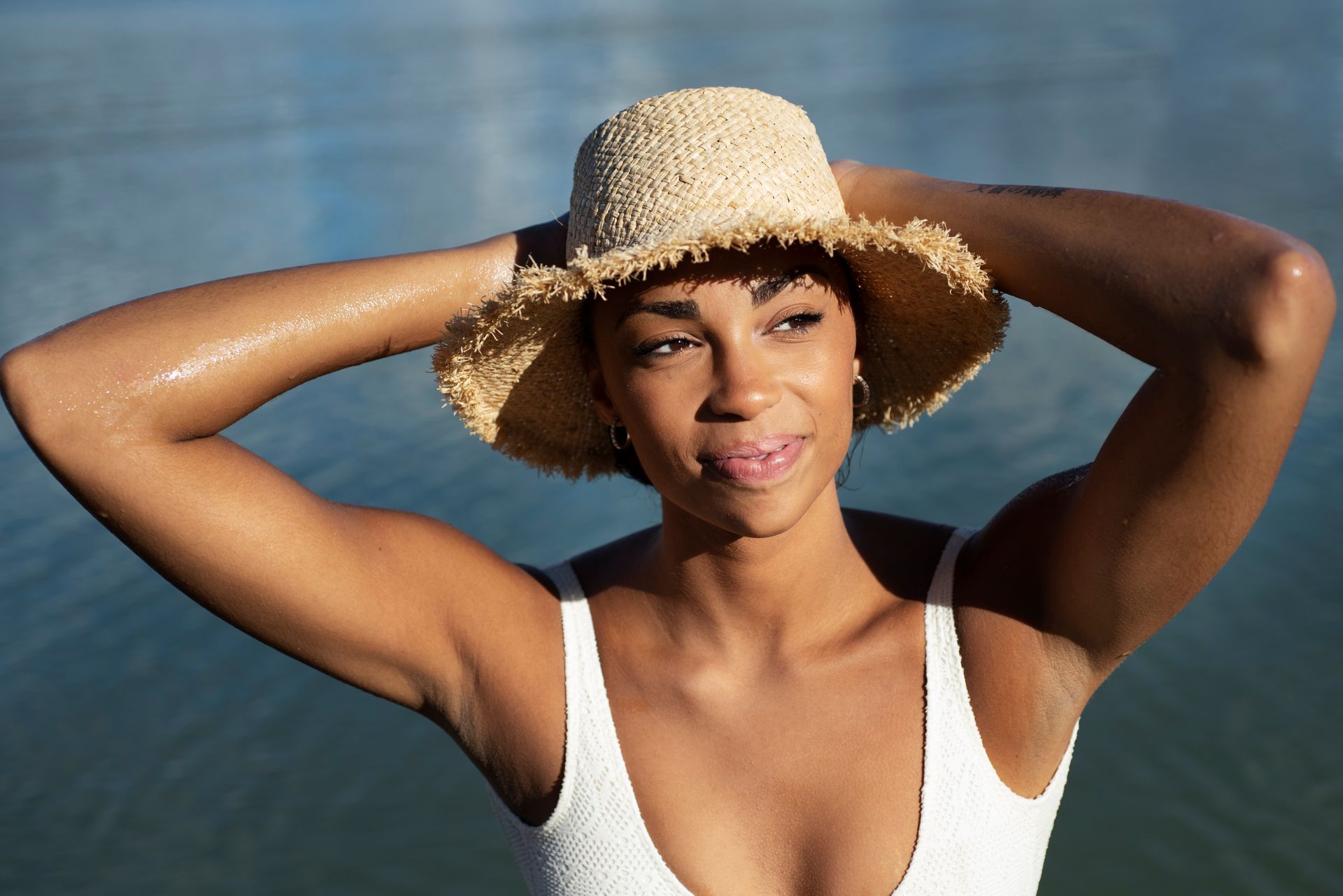 A woman in a white swimsuit and straw hat is standing in front of a body of water.