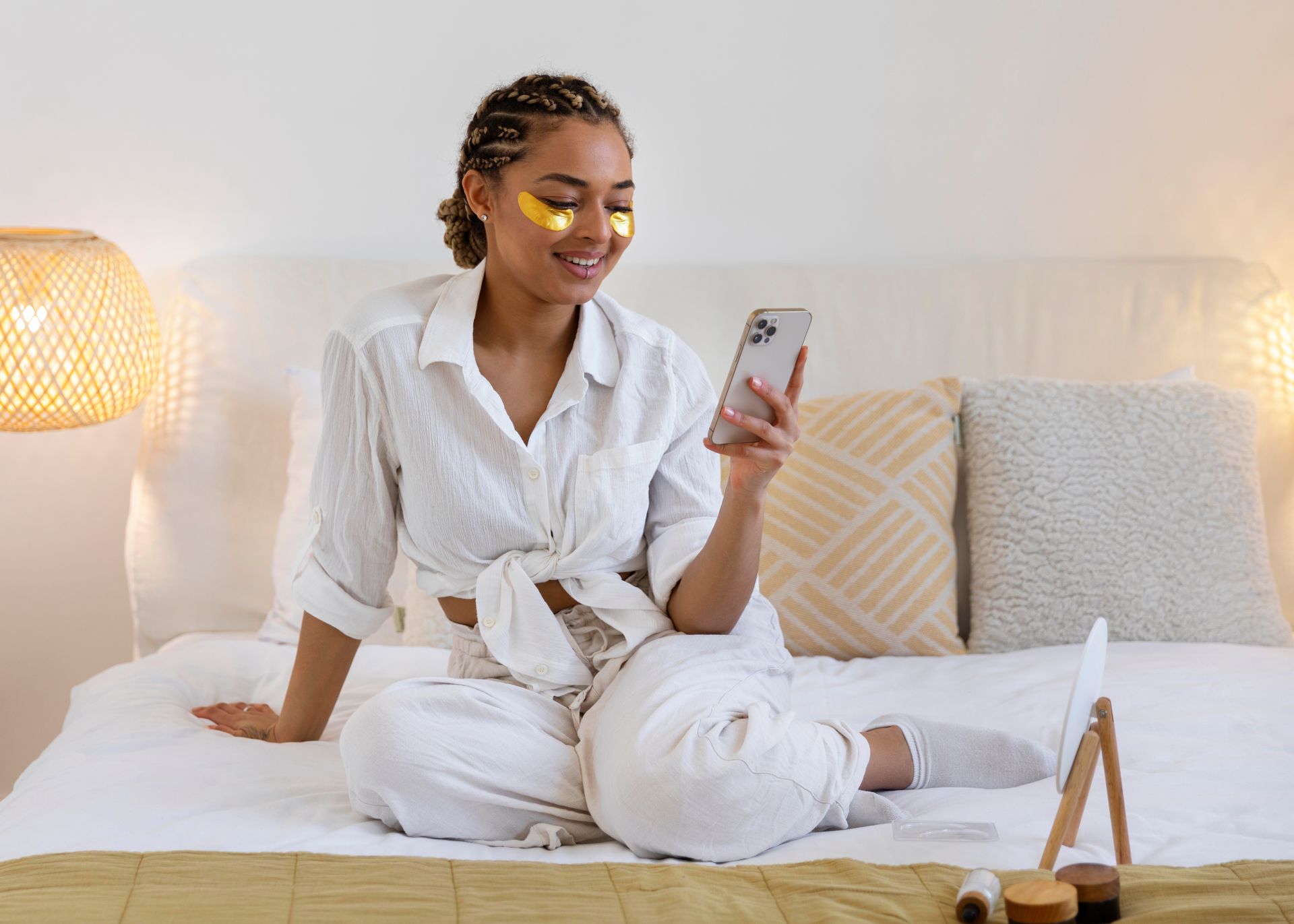 a woman with gold eye patches is sitting on a bed looking at her phone .