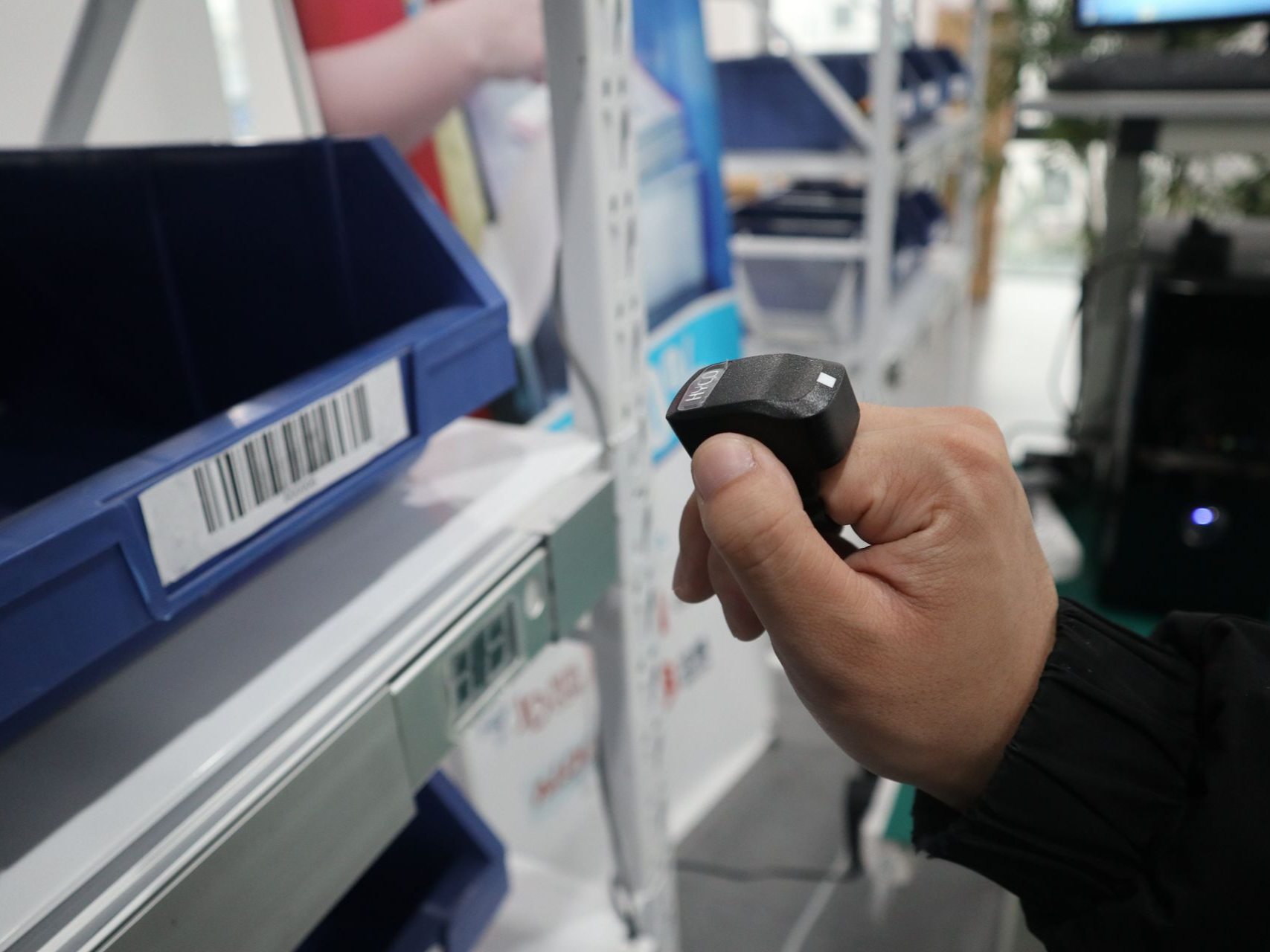 Person scans a barcode on a blue bin with a ring scanner in a warehouse setting.