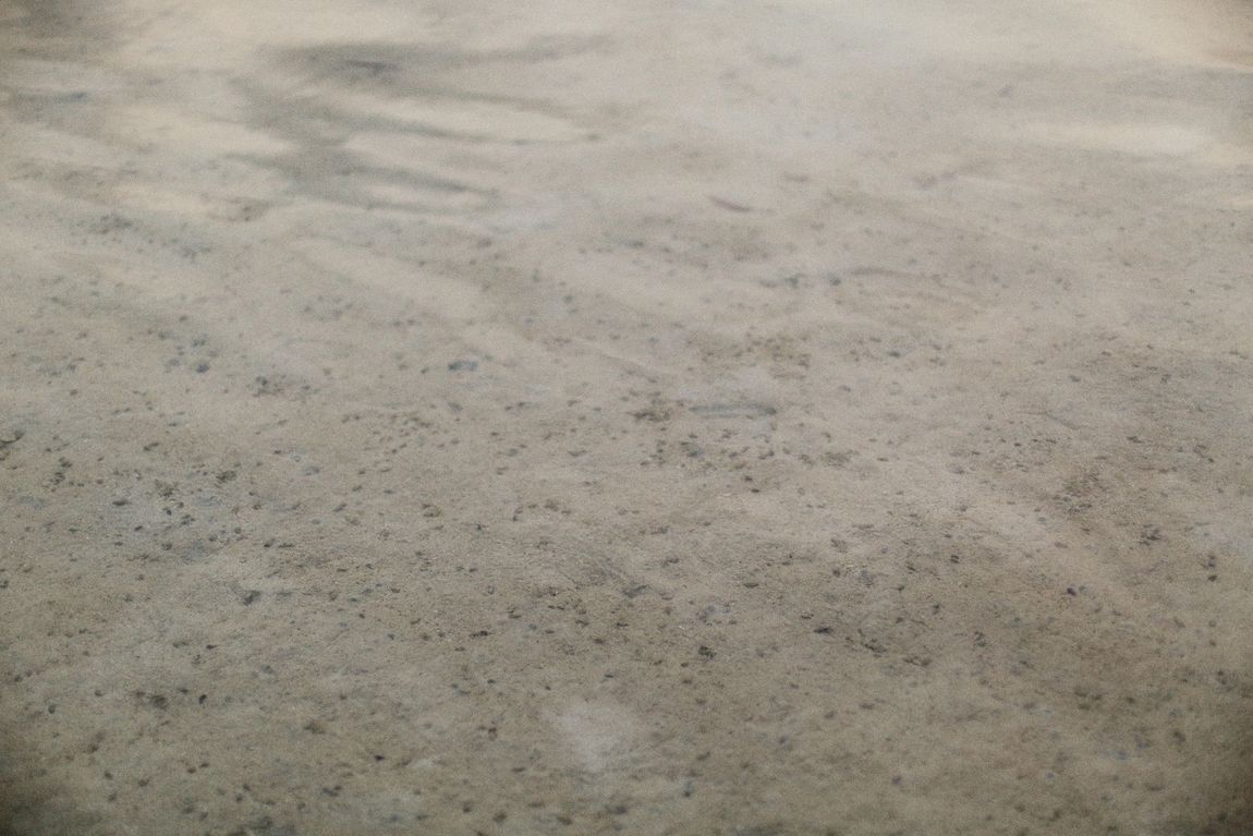 A close-up of a light beige, fine-textured sandy surface with subtle, wavy patterns and small dark specks.