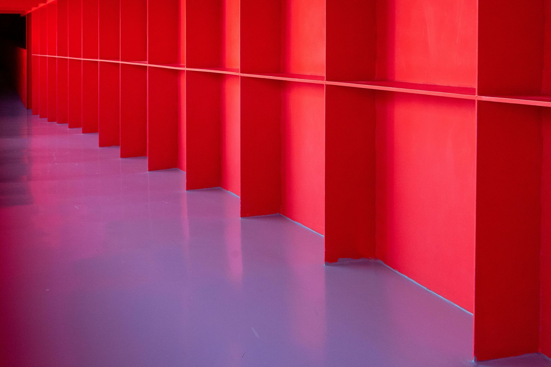 A bright red, angular wall with a horizontal railing runs alongside a smooth, purple floor in a long, empty corridor.