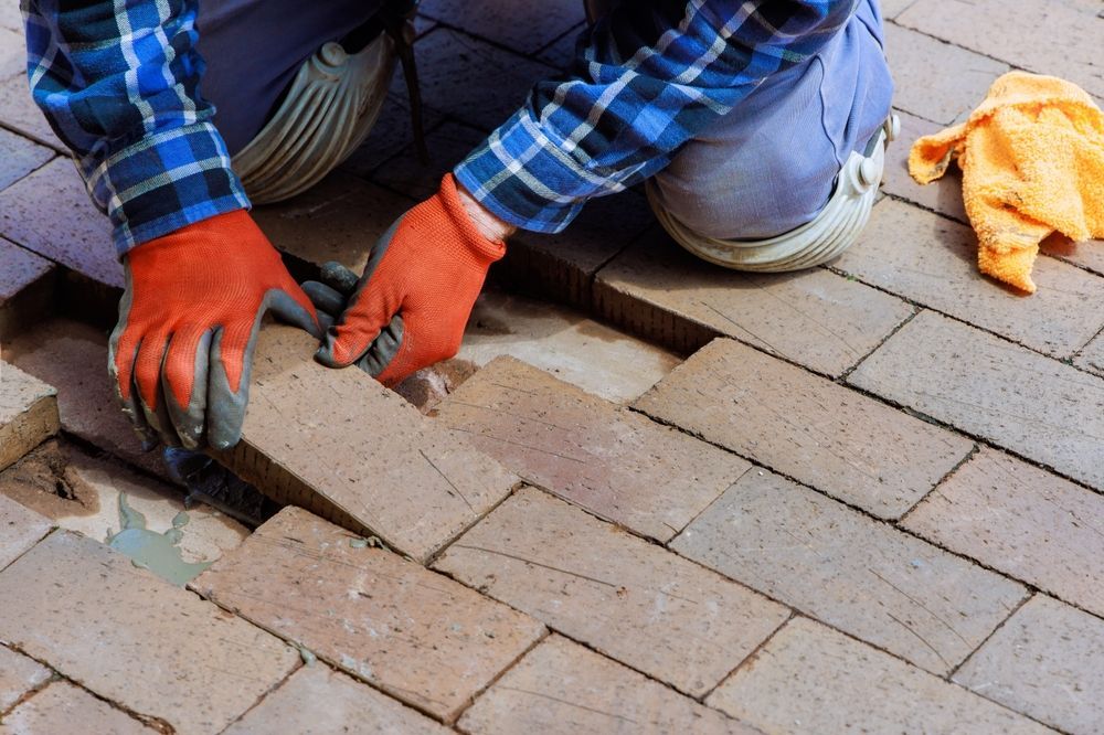 Person in orange gloves laying brick pavers; blue plaid shirt, tan pavers.