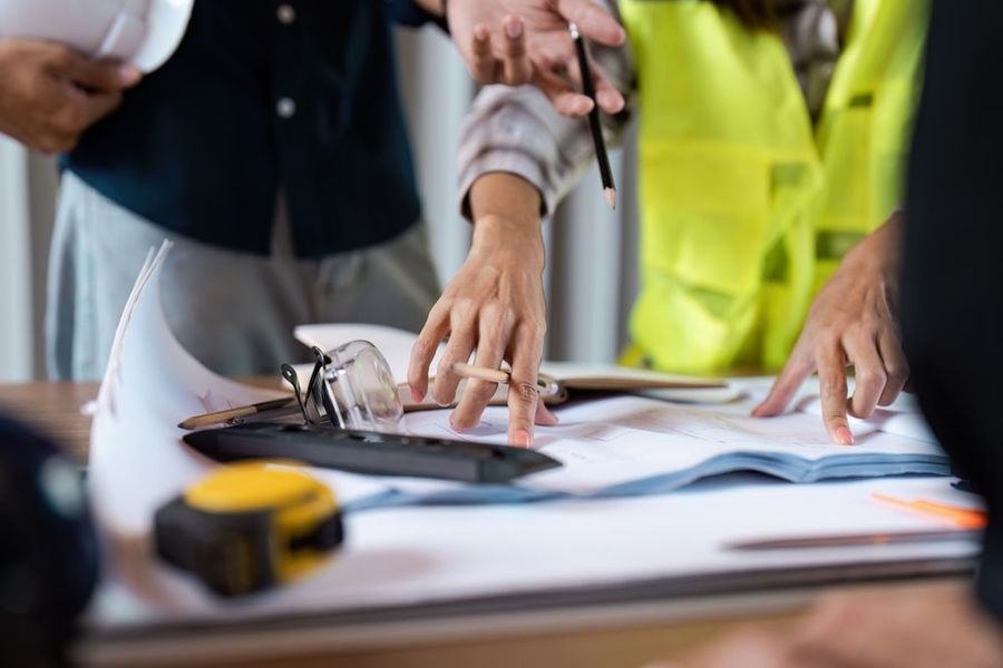 People reviewing blueprints, one holding a hard hat and another wearing a safety vest at a desk.