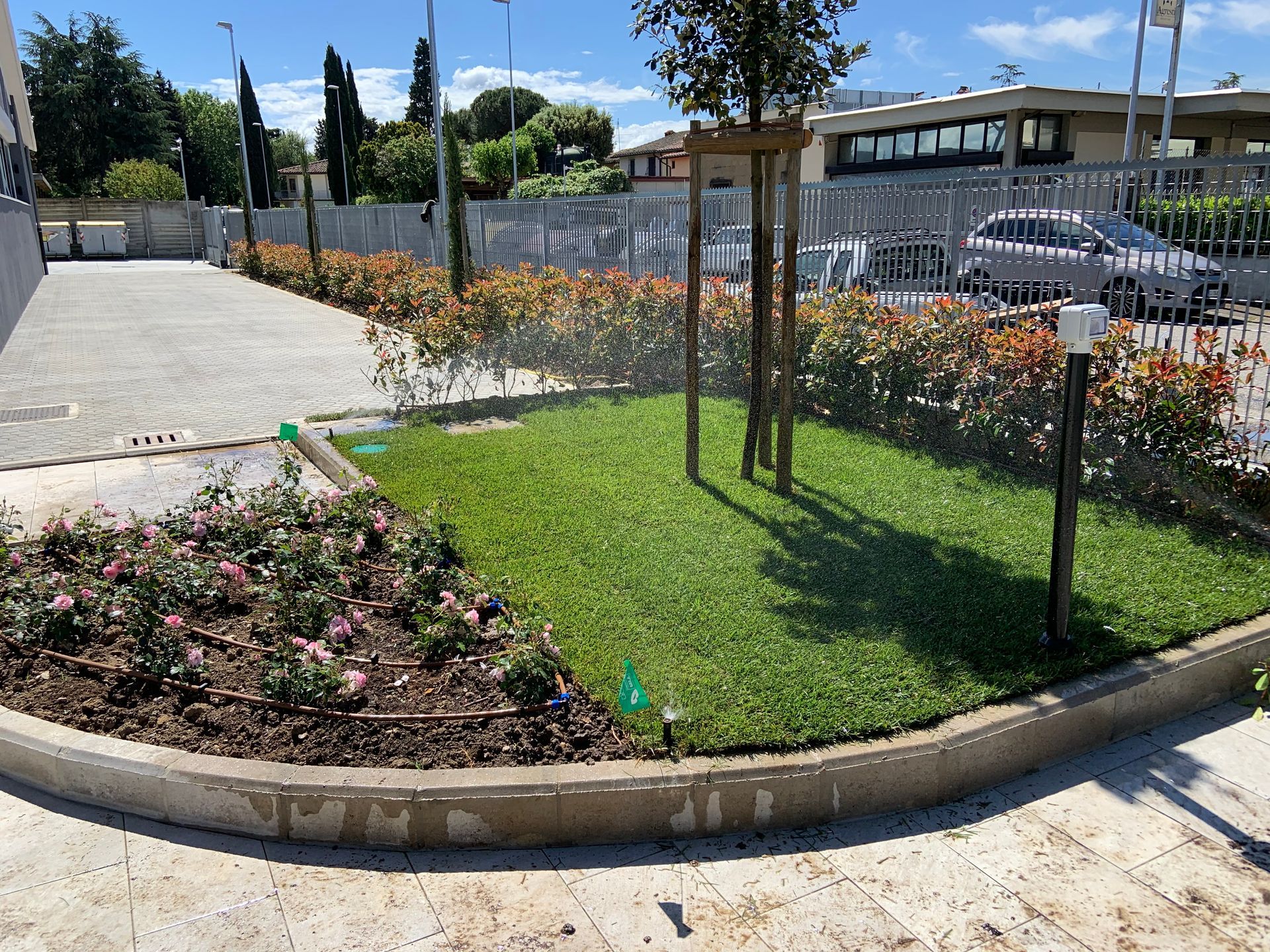 Prato verde rigoglioso e aiuole con irrigatori, delimitato da cemento e da una recinzione metallica, giornata di sole.